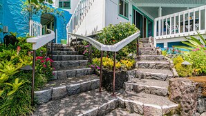 Unos escalones de piedra conducen a un edificio blanco con detalles verdes, rodeado de diversas plantas y flores coloridas.