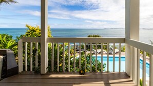 A wooden deck with a white railing overlooking a pool and the ocean.