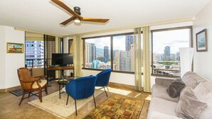Ein modernes Wohnzimmer mit Stadtblick, Deckenventilator, Flachbildfernseher, einer Couch und zwei blauen Sesseln.