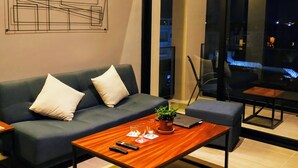 Ein modernes Wohnzimmer mit einem dunkelgrauen Sofa, einem Holztisch und einem Laptop. Ein großes Fenster bietet Blick auf die beleuchtete Stadt bei Nacht.