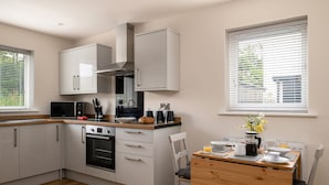 Una cocina moderna con armarios blancos, horno empotrado, microondas y cafetera. Hay una mesa de comedor de madera con sillas y una ventana con persianas.