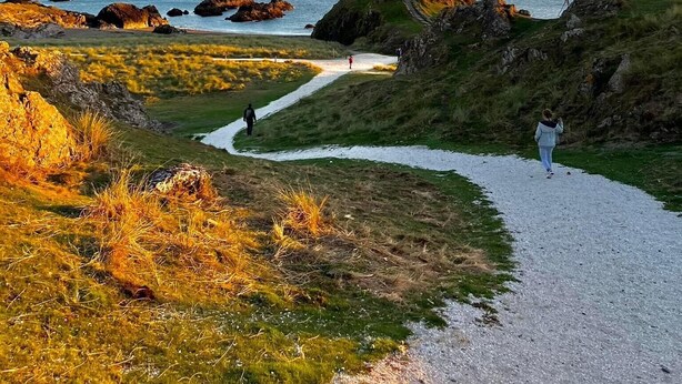 Un sendero costero que lleva a un faro, rodeado de terreno cubierto de hierba y afloramientos rocosos.