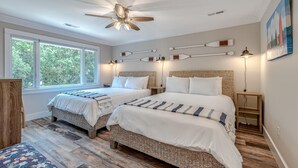 Un dormitorio con dos camas, un ventilador de techo, un ventanal grande y un cuadro en la pared.