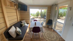 Ein gemütliches Zimmer mit einer Couch, einem Tisch, Stühlen, einem an der Wand befestigten Fernseher und Blick ins Freie durch eine Glastür.