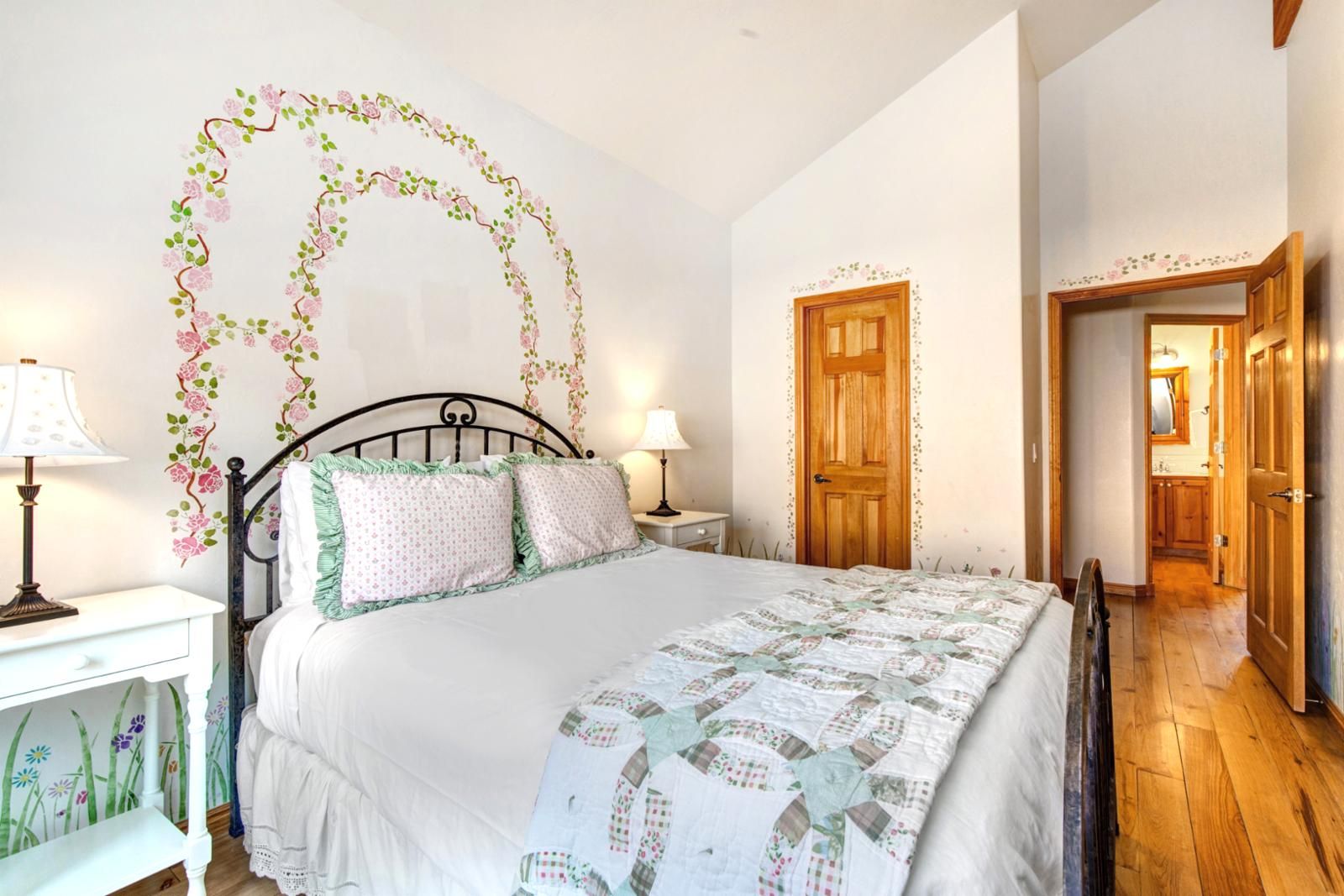 A bedroom with a bed, two bedside tables, a nightstand, a wooden door, and a hallway leading to a bathroom.