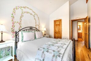 Un dormitorio con una cama, dos mesitas de noche, un tocador, una puerta de madera y un pasillo que conduce a un baño.