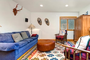 Sala de estar com sofá azul, um aparador de madeira, tapete estampado e decorações nas paredes, incluindo um crânio e duas máscaras.