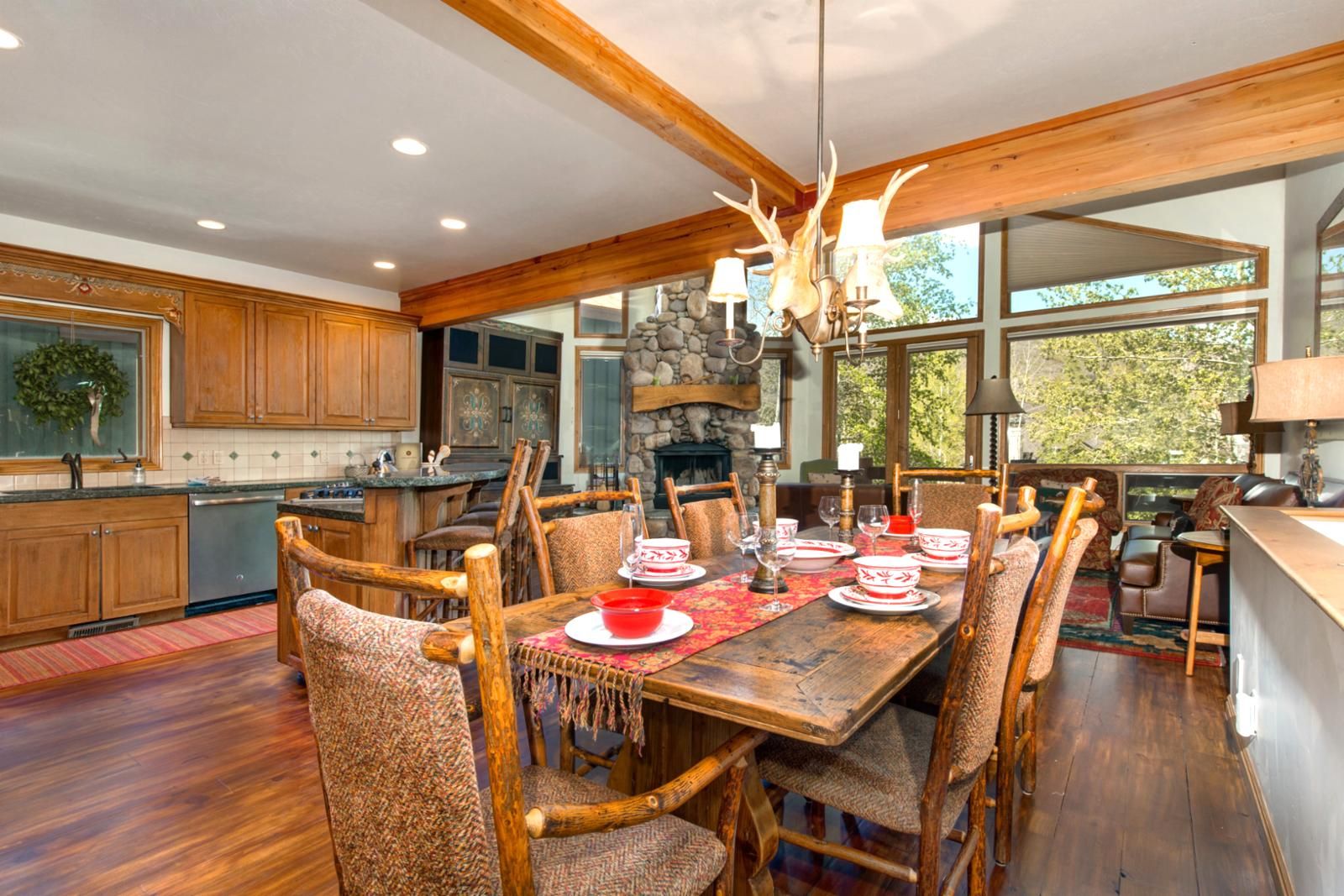 A spacious dining area with a wooden table set for six, surrounded by rustic chairs. A stone fireplace and large windows provide natural light.