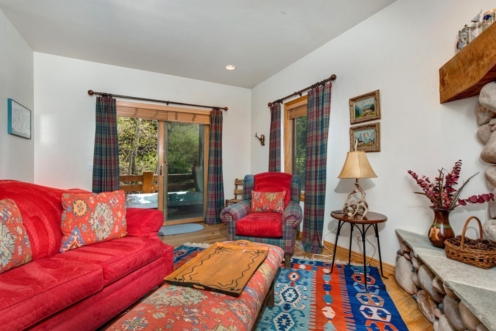 A living room with a red sofa, a patterned ottoman, a plaid armchair, and a small side table with a lamp.