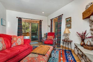 Una sala de estar con un sofá rojo, un reposapiés estampado, un sillón escocés y una mesita auxiliar con lámpara.