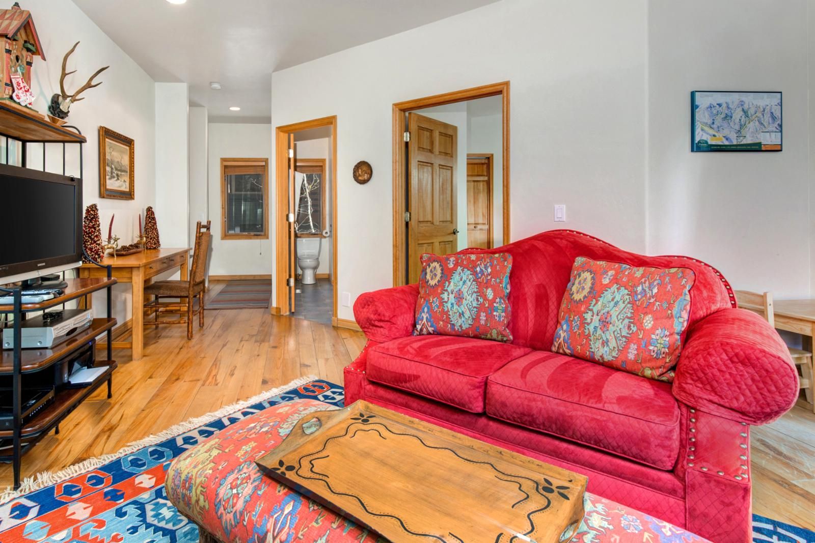 A living room with a red sofa, wooden coffee table, and patterned rug. There is a television on a wooden stand, a dining table with chairs, and a door leading to another room.