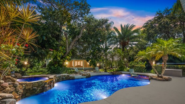 A pool with a curved edge, surrounded by rocks and plants, with a building in the background. - Tropical Resort Style Retreat w/ Pool House 4BR, 3 Bath (Stuart)