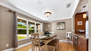 A spacious dining area with a wooden table and chairs, a kitchen with wooden cabinets, and a large window with curtains. - Tropical Resort Style Retreat w/ Pool House 4BR, 3 Bath (Stuart)