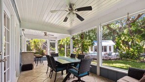A covered patio with a dining table and chairs, a ceiling fan, and large windows overlooking a garden and pool area. - Tropical Resort Style Retreat w/ Pool House 4BR, 3 Bath (Stuart)
