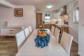 Un coin repas avec une table blanche, une nappe bleue et un panier de fruits. On aperçoit, au fond, une cuisine avec des meubles en bois et une fenêtre.