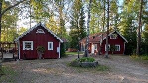 Mökkiympäristö, jossa on kaksi punaiseksi maalattua puumökkiä, katettu oleskelualue ja sorapolkua.