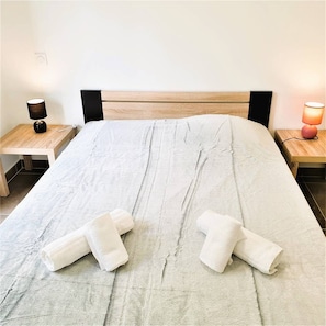 Cama bem arrumada com um edredom claro, dois toalhas enroladas sobre a cama e duas mesas de cabeceira com abajures.