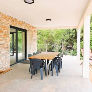 Eine überdachte Terrasse mit einem Holztisch und -stühlen, eine Glastür, die zu einem Außenbereich führt, und ein Metallzaun.