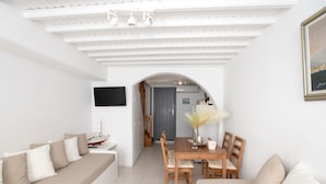 A modern living room with a sofa, dining table, and chairs. There is a TV mounted on the wall and a framed picture above the table. - Alessios Mykonian Downtown Houses (Mykonos)