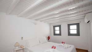 A white bedroom with a bed, bedside table, lamp, and two windows. - Alessios Mykonian Downtown Houses (Mykonos)