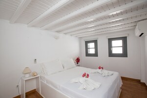 Un dormitorio blanco con cama, mesita de noche, lámpara y dos ventanas.