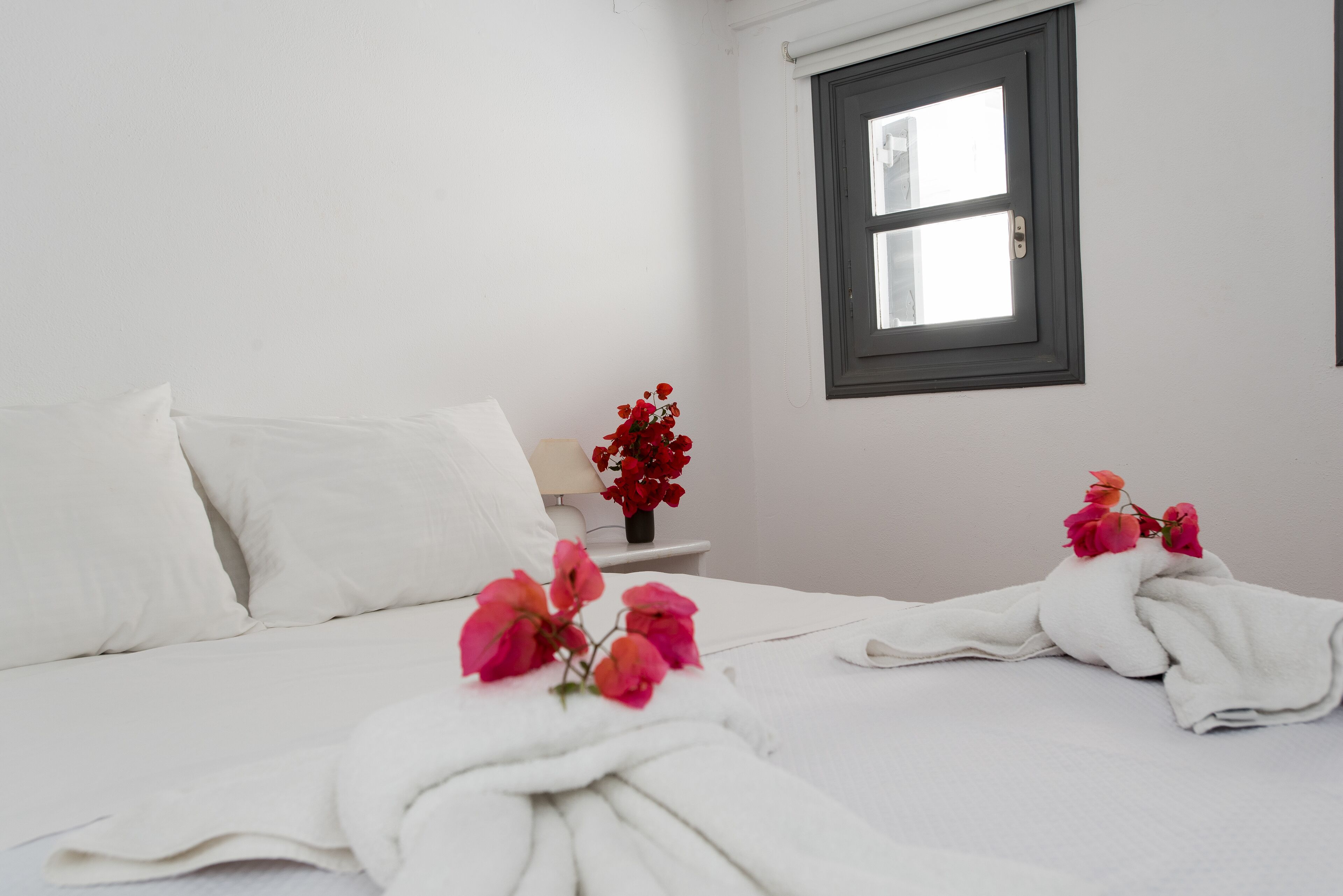 A hotel room with a bed, a nightstand, a window, and a vase of flowers.
