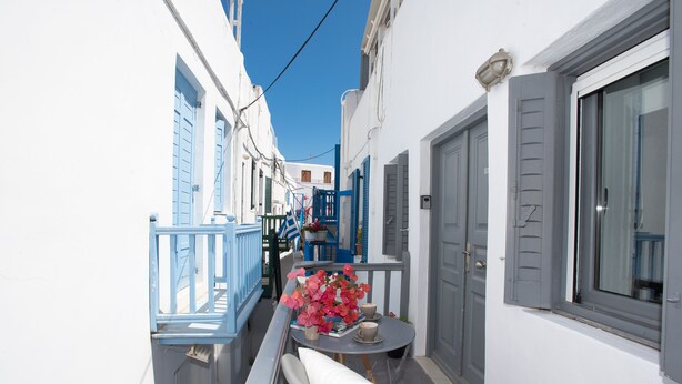 A narrow street with white buildings, blue shutters, and a table with a vase of flowers. - Alessios Mykonian Downtown Houses (Mykonos)