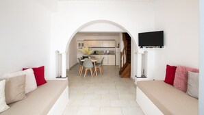 A modern living room with a sofa, a dining area with chairs, and a kitchen in the background. - Alessios Mykonian Downtown Houses (Mykonos)