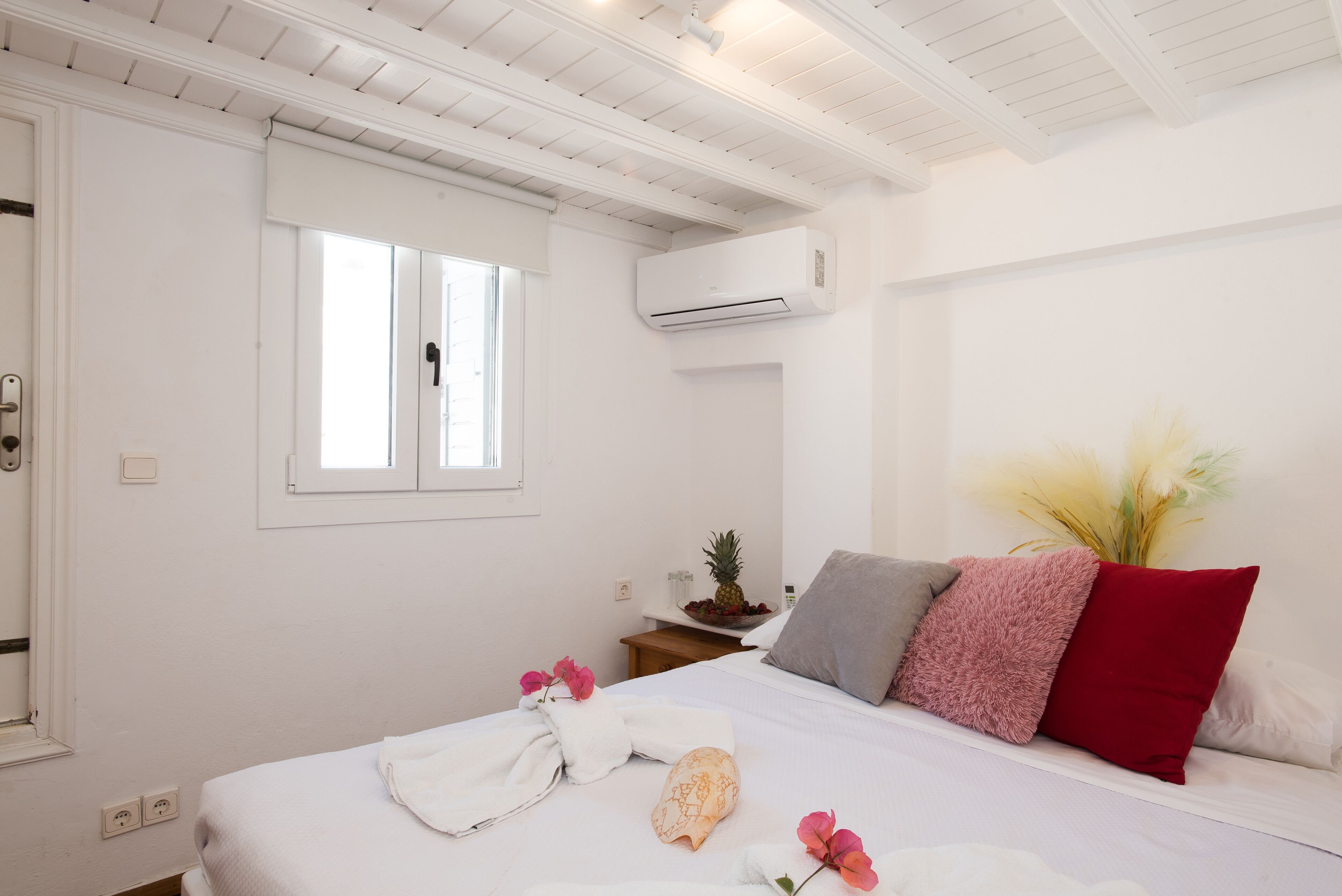 A bedroom with a bed, a window, an air conditioner, a nightstand with a plant, and a wall-mounted shelf with a pineapple.