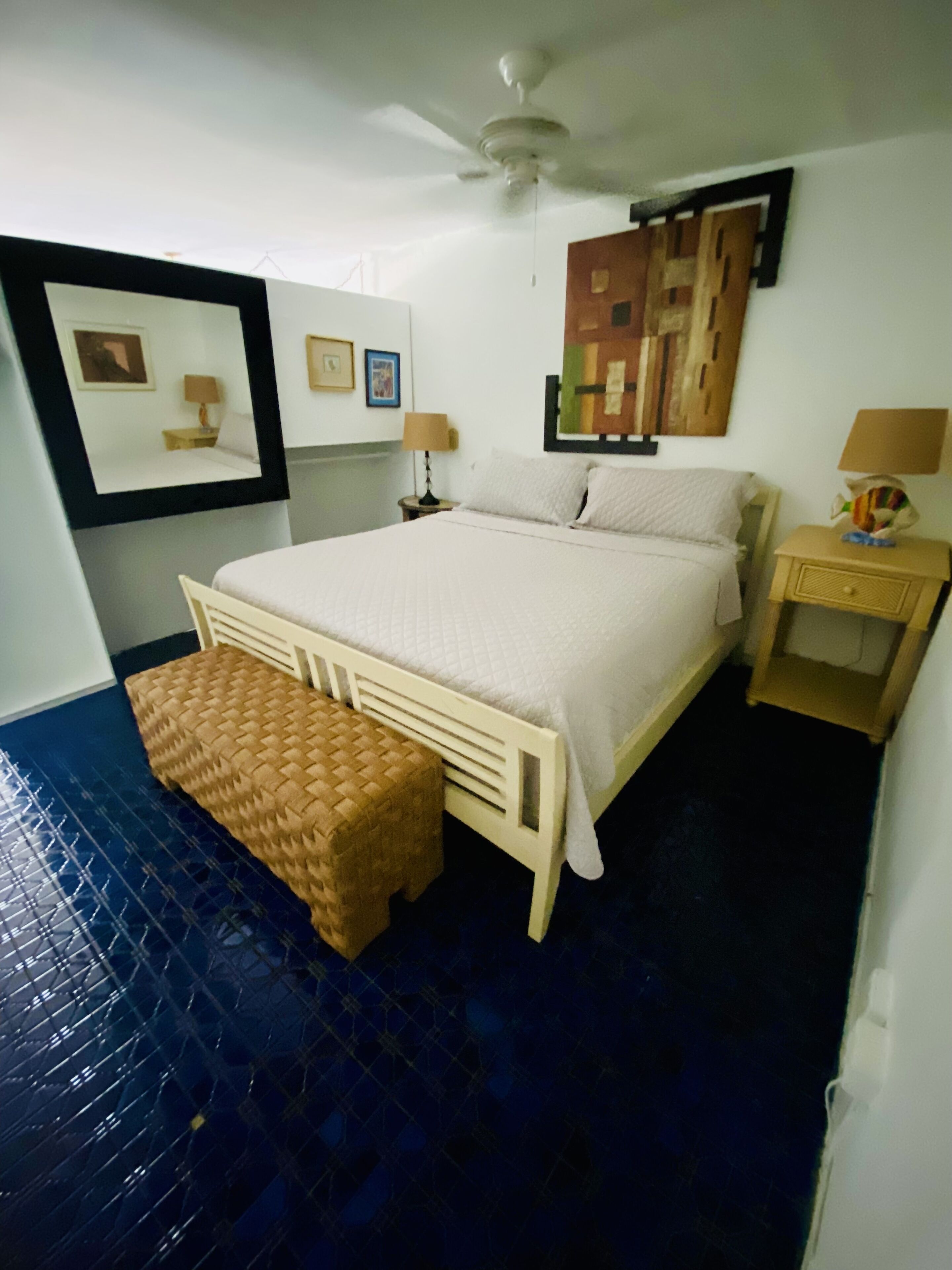 A bedroom with a bed, a nightstand, a lamp, a ceiling fan, a wall-mounted artwork, and a woven bench.