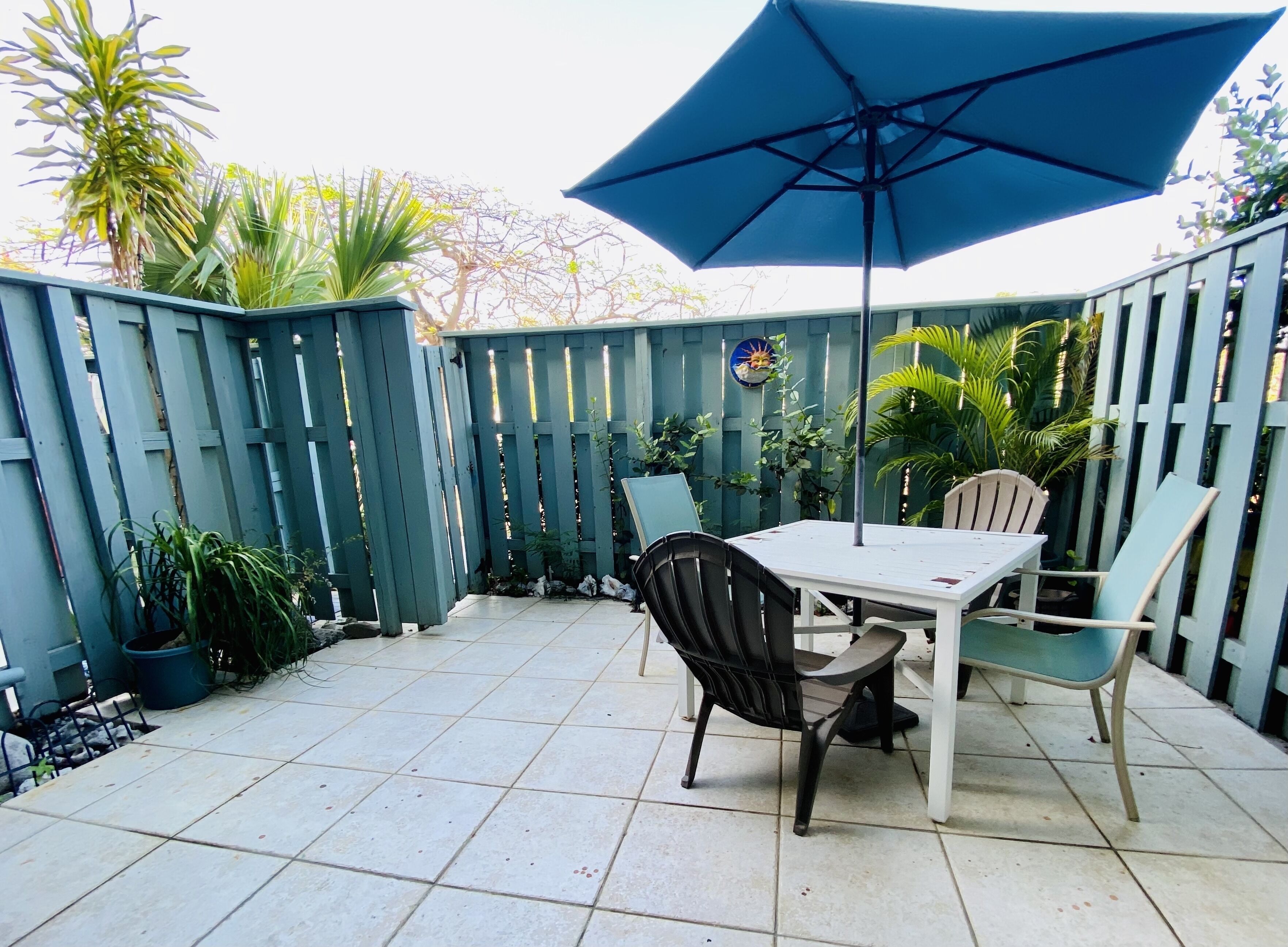 A patio with a table and chairs, a blue umbrella, and a wooden fence.