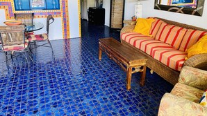 A room with a blue tiled floor, a wicker sofa with striped cushions, a wooden coffee table, a wicker chair, and a small round table with chairs.