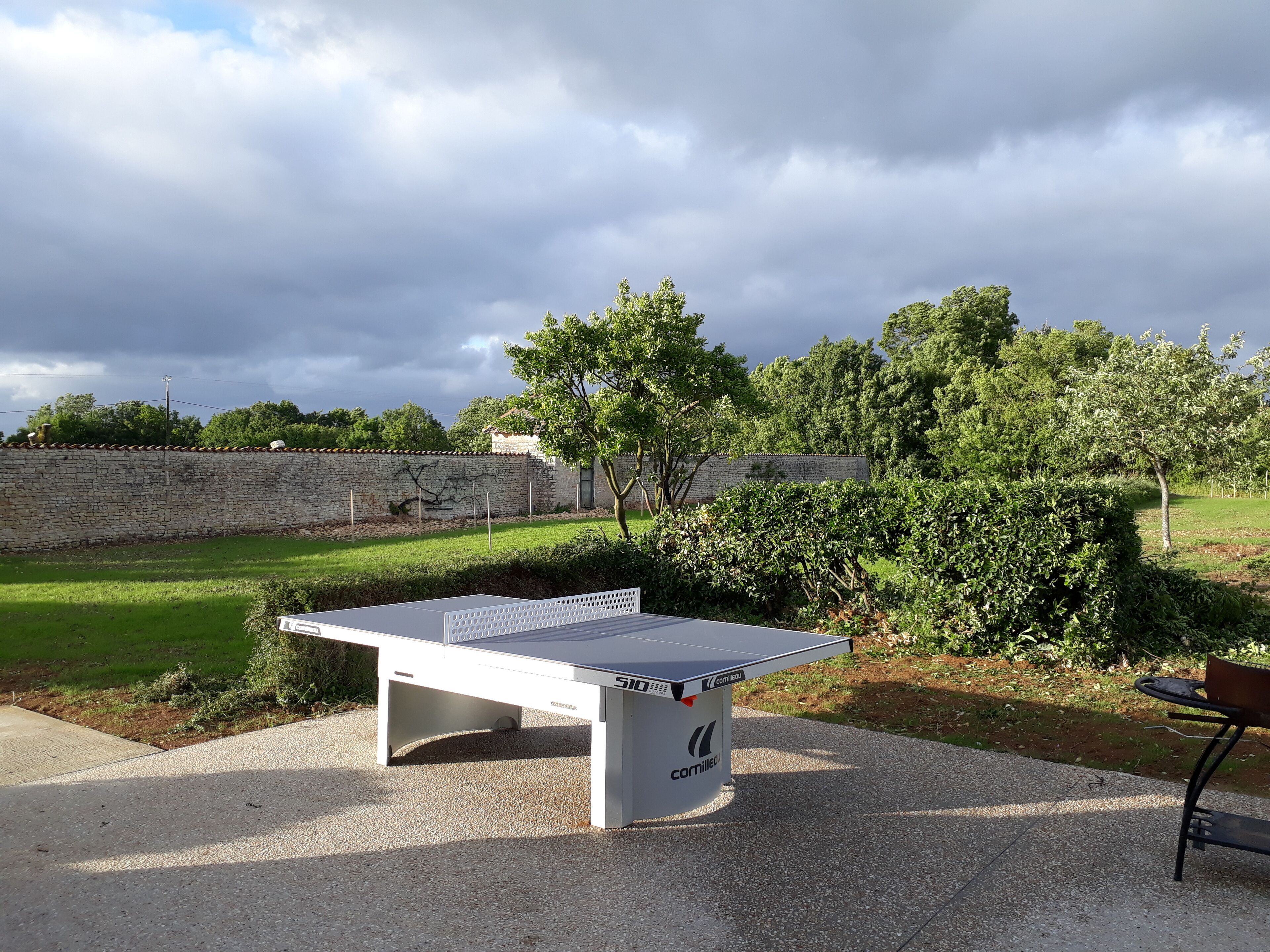 A ping pong table in an outdoor area with a stone wall, trees, and a grassy field.