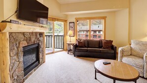 A living room with a stone fireplace, a flat-screen TV mounted above it, a brown leather sofa, a patterned armchair, and a wooden coffee table. - Top-Floor Condo with Stunning Views, Hot Tub, and Vaulted Ceilings - CS425 (Copper Mountain)