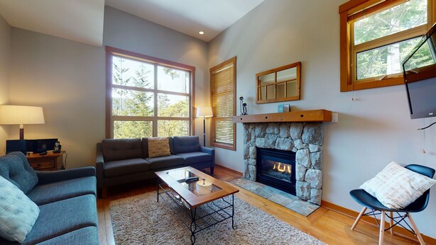 A living room with a stone fireplace, a sofa, a coffee table, and a TV mounted on the wall. - 3 Bedroom plus Den / 2.5 Bath Mountain Star Townhome, Private Hot Tub, Quiet Slope Side location (Whistler)
