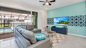 A modern living room with a grey sofa, a coffee table, a TV, and a ceiling fan. - Ultimate 5 Bedroom Windsor Island Resort Pool Town home (Davenport)