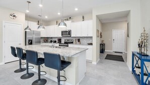 A modern kitchen with white cabinets, a granite countertop, and stainless steel appliances. There are four bar stools and pendant lights above the island. - Ultimate 5 Bedroom Windsor Island Resort Pool Town home (Davenport)