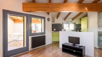 A modern living room with a television, wooden beams, a sliding glass door, and a view into the kitchen.