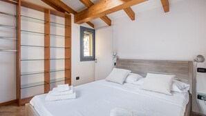 A bedroom with a wooden ceiling, a bed with white bedding, a glass shelving unit, and a small window.