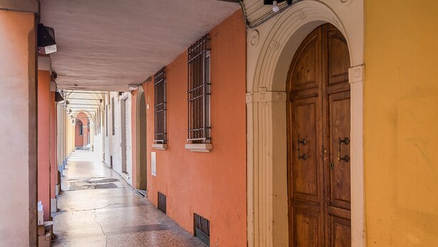 A corridor with a wooden door and windows, leading to a building with a yellow wall.