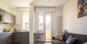 A modern apartment with a kitchen, a dining area, and a living room. There is a balcony with a view of buildings.