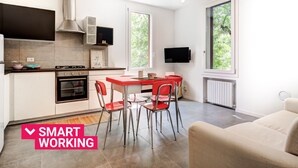 A modern kitchen with a dining area, a TV mounted on the wall, and a large window providing a view of greenery.