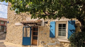 Un edificio de piedra con puerta y contraventanas azules, una pequeña mesa con sillas y una planta en maceta.