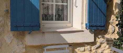 A stone building with a window and blue shutters, a small table, and a chair.