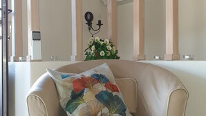 Un sillón beige con un cojín floral, una lámpara de pared y una planta pequeña en una estantería.