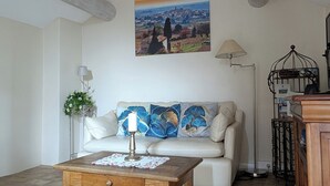 Un salón con sofá blanco, una mesa de centro de madera con un mantelillo, un cuadro de un paisaje, una lámpara de pie y una jaula de pájaros.