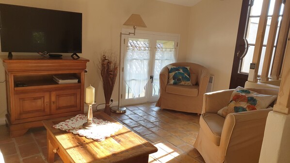 A living room with a television on a wooden cabinet, a table with a candle, a chair with a floral cushion, and a door with a curtain.