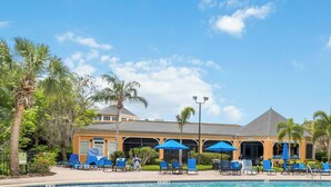 A pool area with lounge chairs and umbrellas, surrounded by palm trees and a building with a grey roof.