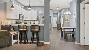 A modern kitchen with white cabinets, a microwave, and a sink. There are two bar stools and a dining table with chairs. The room has a gray wall and a wooden floor.