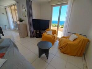 A modern living room with a flat-screen TV, two yellow-covered sofas, a small round table, and a sliding glass door leading to a balcony with a view of the sea. - Magnificent House for 8–12 People in Porto Vecchio with Ocean Views of Pallombaggia and Santa Giulia (Porto-Vecchio)